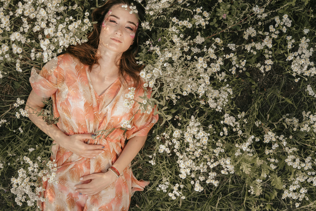 Frau liegt in einem Blumenfeld auf dem Rücken und hält beide Hände auf ihrem Bauch.