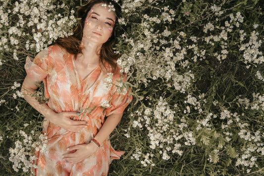 Frau liegt in einem Blumenfeld auf dem Rücken und hält beide Hände auf ihrem Bauch.